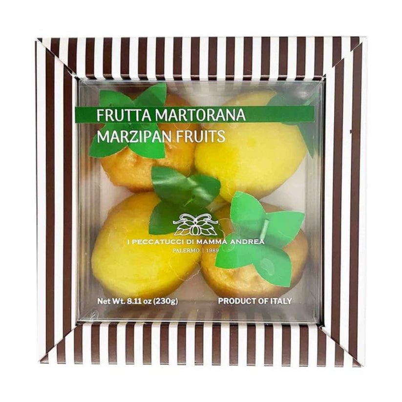 A boxed set of marzipan fruits, including yellow almond paste fruits and green leaf-shaped marzipan decorations, presented in a clear square package with a brown-and-white striped border and a green label reading 'Frutta Martorana Marzipan Fruits.'.
